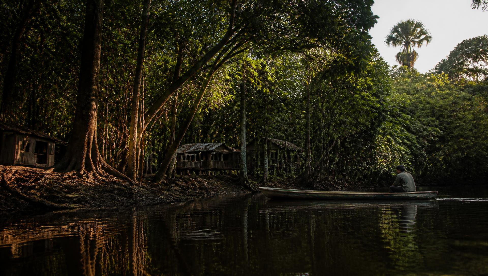 Igarapé de águas escuras no Tarumã Mirim, com floresta densa nas margens, casas ribeirinhas de madeira e uma canoa com um morador ao entardecer.